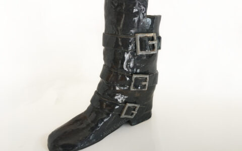 A photo of a ceramic black women's boot with three black straps and grey buckles against a white background.