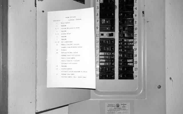 A black and white photo of a circuit breaker panel with each breaker labelled with a number written in calligraphic style. A half-sheet of paper is taped to the inside of the door with typewriter font detailing what circuits in the house correspond to a given number.